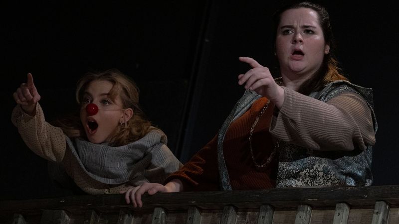 Two characters in red clown noses leaning over the gallery rail, pointing and mid-shout