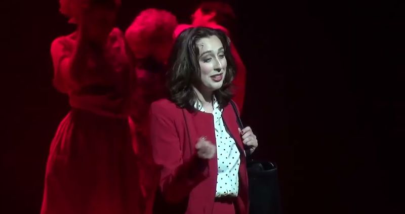 A woman in a red jacket downstage in a warm red wash, two figures behind her in silhouette