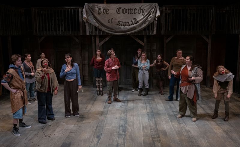 The full company of The Comedy of Errors beneath a hand-painted banner