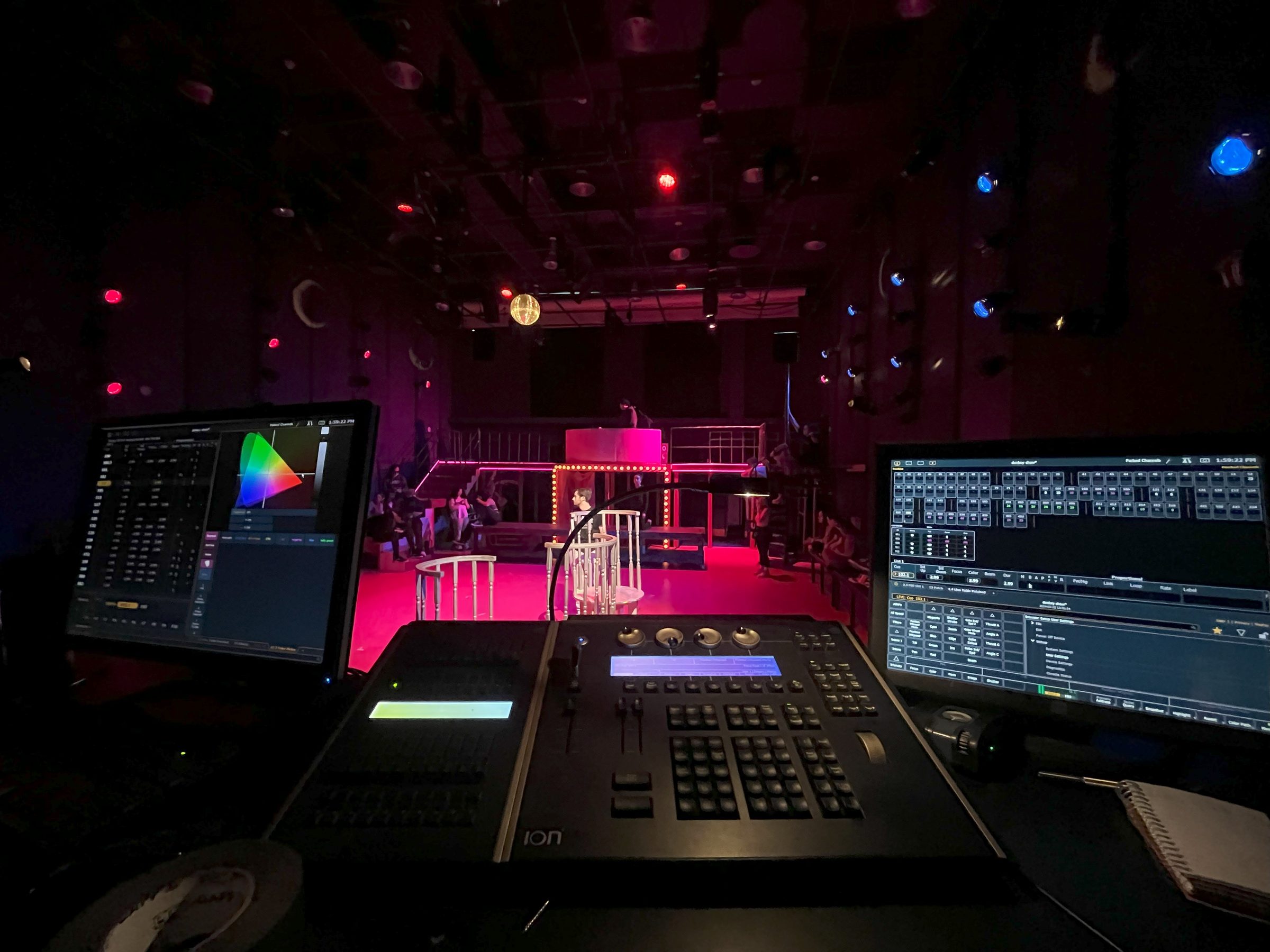 A lighting desk and its monitors in a booth, the room washed red behind the screens